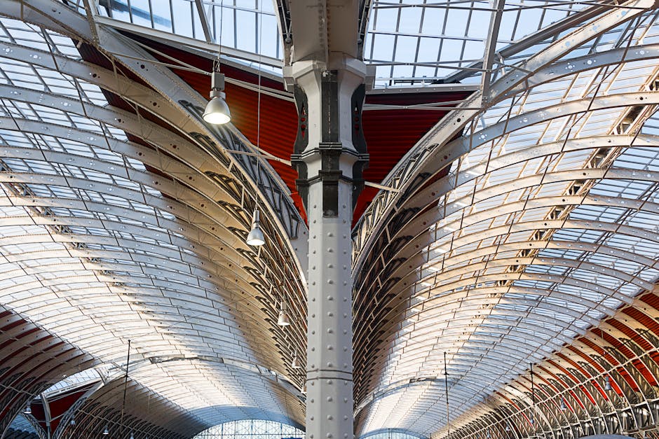 A close-up view of the interior roof structure of a modern building, featuring a central vertical steel support column with rivets, surrounded by curved, overlapping metal panels and glass panels that
