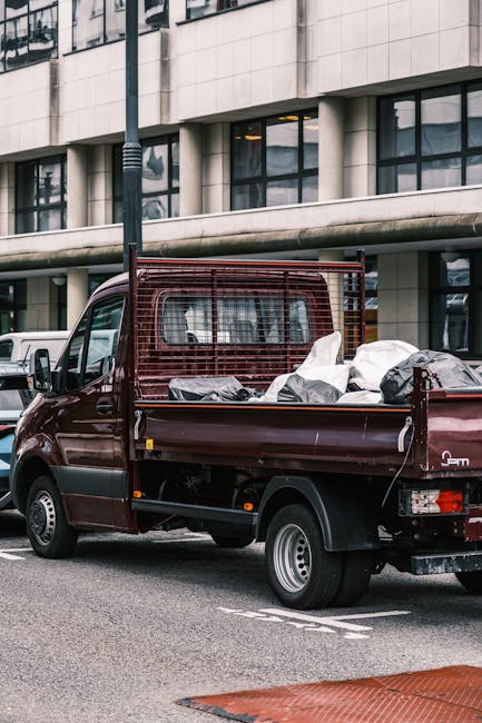 A burgundy flatbed utility truck parked on an urban street in front of a modern office building with large glass windows and a concrete facade. The truck's cargo area is filled with various types of w