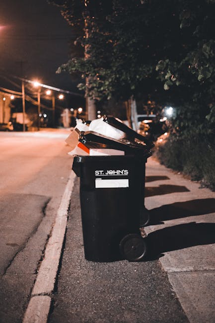 A large green wheeled rubbish bin positioned outdoors on a paved surface against a dark brick wall. The bin is filled with multiple red and black flexible garden hose reels, featuring labels and brand